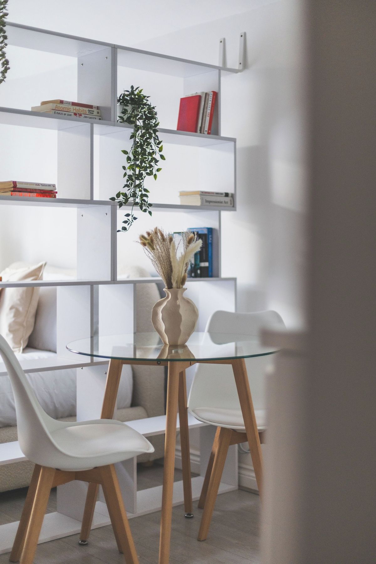 A modern living room with a glass dining table, white chairs, a sofa, and a bookshelf.