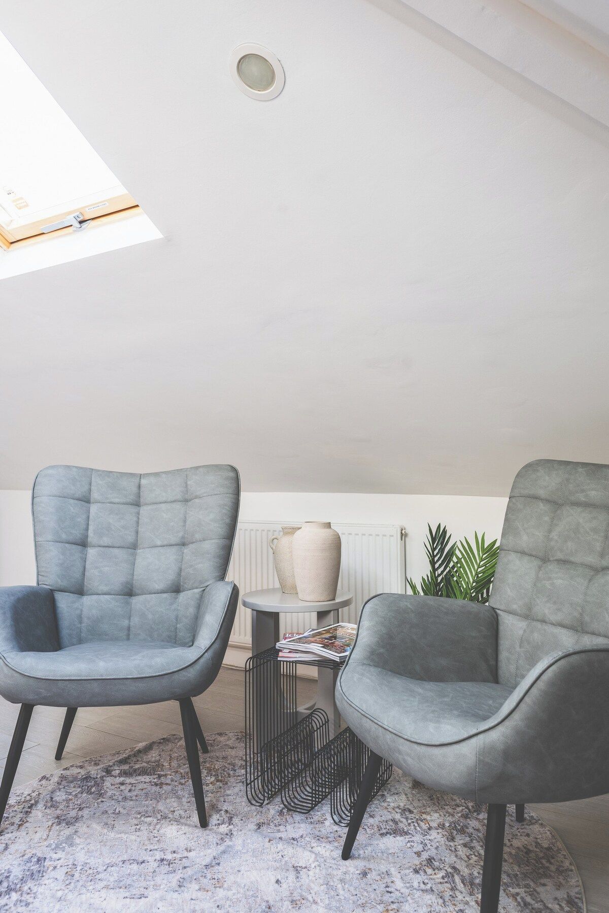A modern living room with two grey upholstered chairs, a small round table with a lamp, and a radiator in the background.