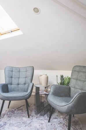 Ein modernes Wohnzimmer mit zwei grauen Polstersesseln, einem kleinen runden Tisch mit einer Lampe und einem Heizkörper im Hintergrund.