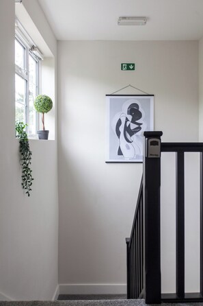 A hallway with a framed abstract artwork, a potted plant, and a black railing. - Chic and Classy 2BR Penthouse w/ Parking, 6 Guests (Greater London)