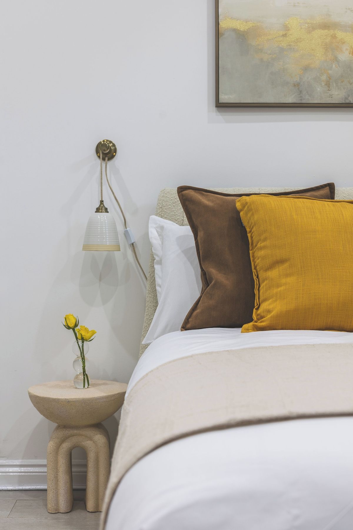 Een keurig opgemaakt slaapkamer met een bed, een nachtkastje, een lamp en een vaas met bloemen.