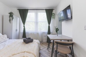 Une chambre à coucher avec un lit, un bureau et une chaise. Il y a une fenêtre avec des rideaux, un radiateur et un téléviseur fixé au mur.