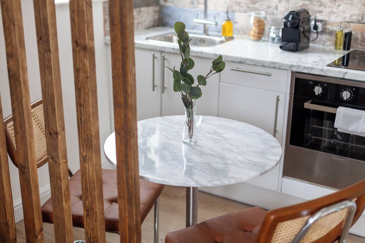 Un tavolo rotondo con un vaso di foglie verdi, una sedia di legno e una cucina sullo sfondo.