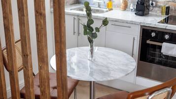 Un tavolo rotondo con un vaso di foglie verdi, una sedia di legno e una cucina sullo sfondo.