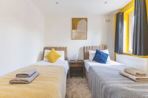 Hotellihuone, jossa on kaksi sänkyä, yöpöytä, ikkuna verhoilla ja seinälle kiinnitetty taideteos.