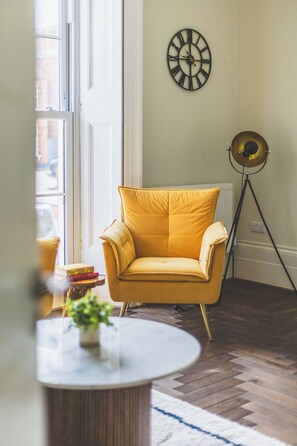 A room with a yellow armchair, a round table with a plant, a floor lamp, and a wall clock. - 3 bed | Sleeps 7 | Private Parking |Great Location (Greater London)
