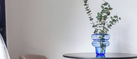 A room with a round table, two chairs, a blue vase with a plant, a framed abstract painting, and a white curtain.