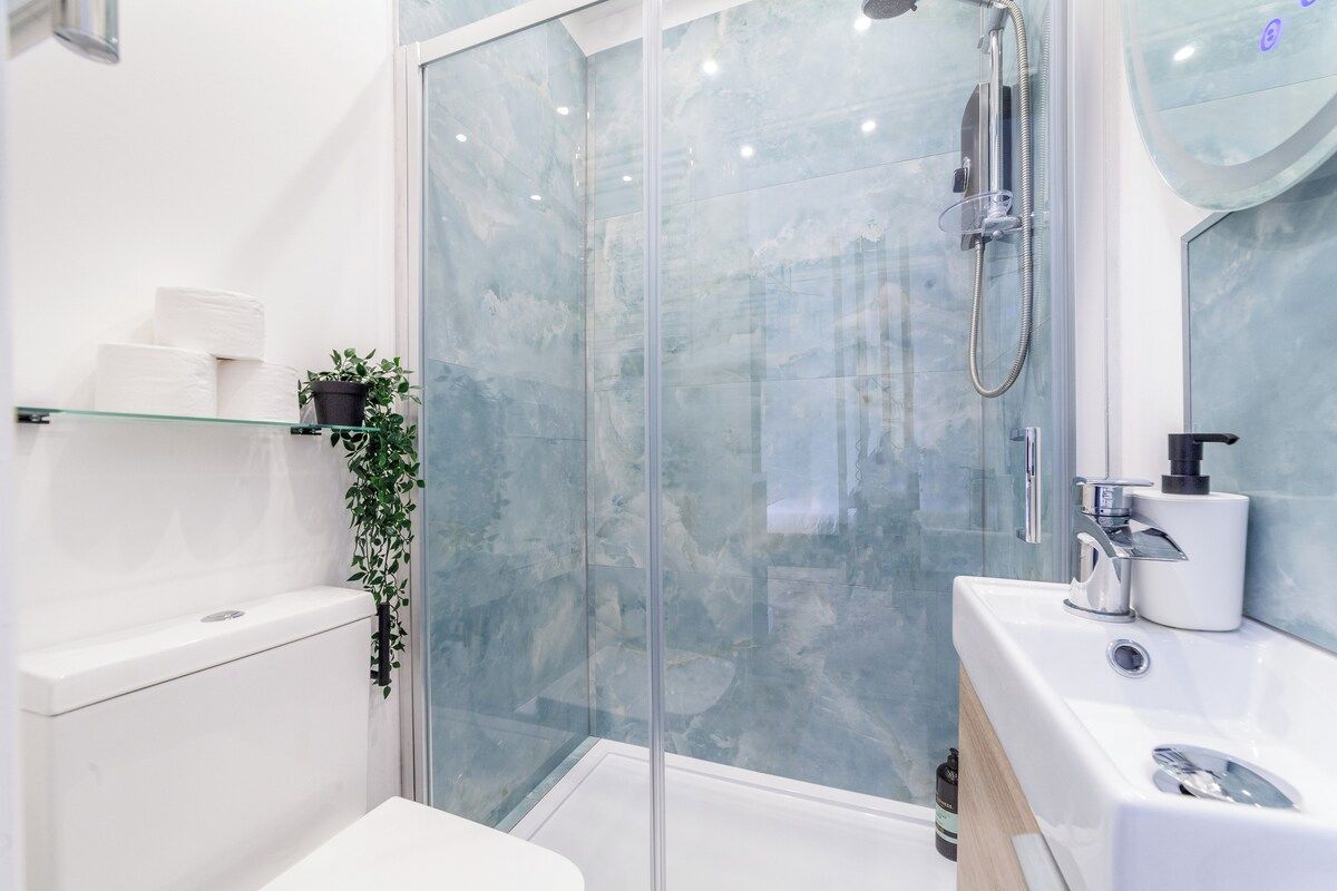Un baño moderno con cabina de ducha de vidrio, inodoro blanco, lavamanos con espejo y una pequeña planta en maceta.
