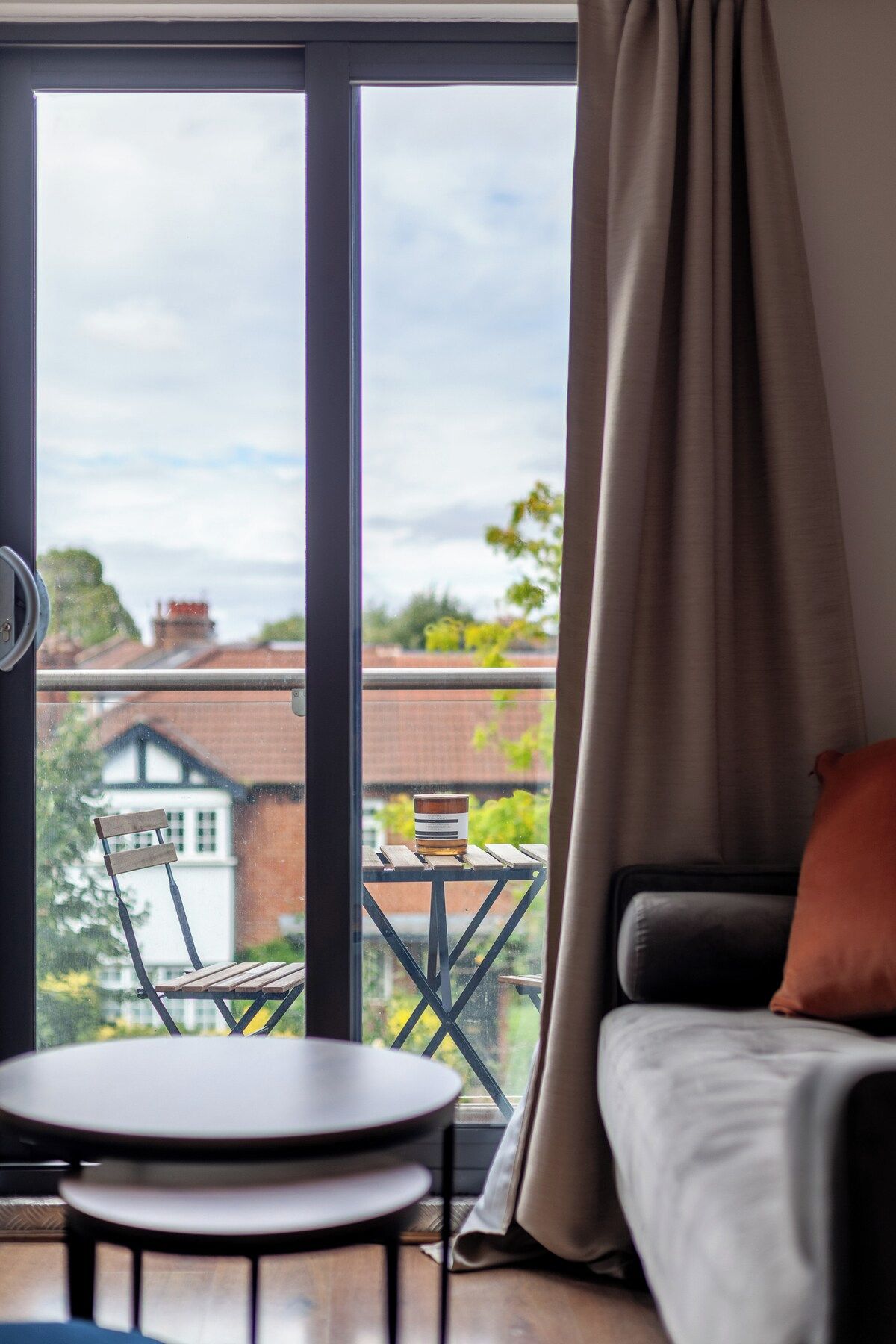 A room with a view of a house through a large window, a small table, a chair, and a sofa with a cushion.