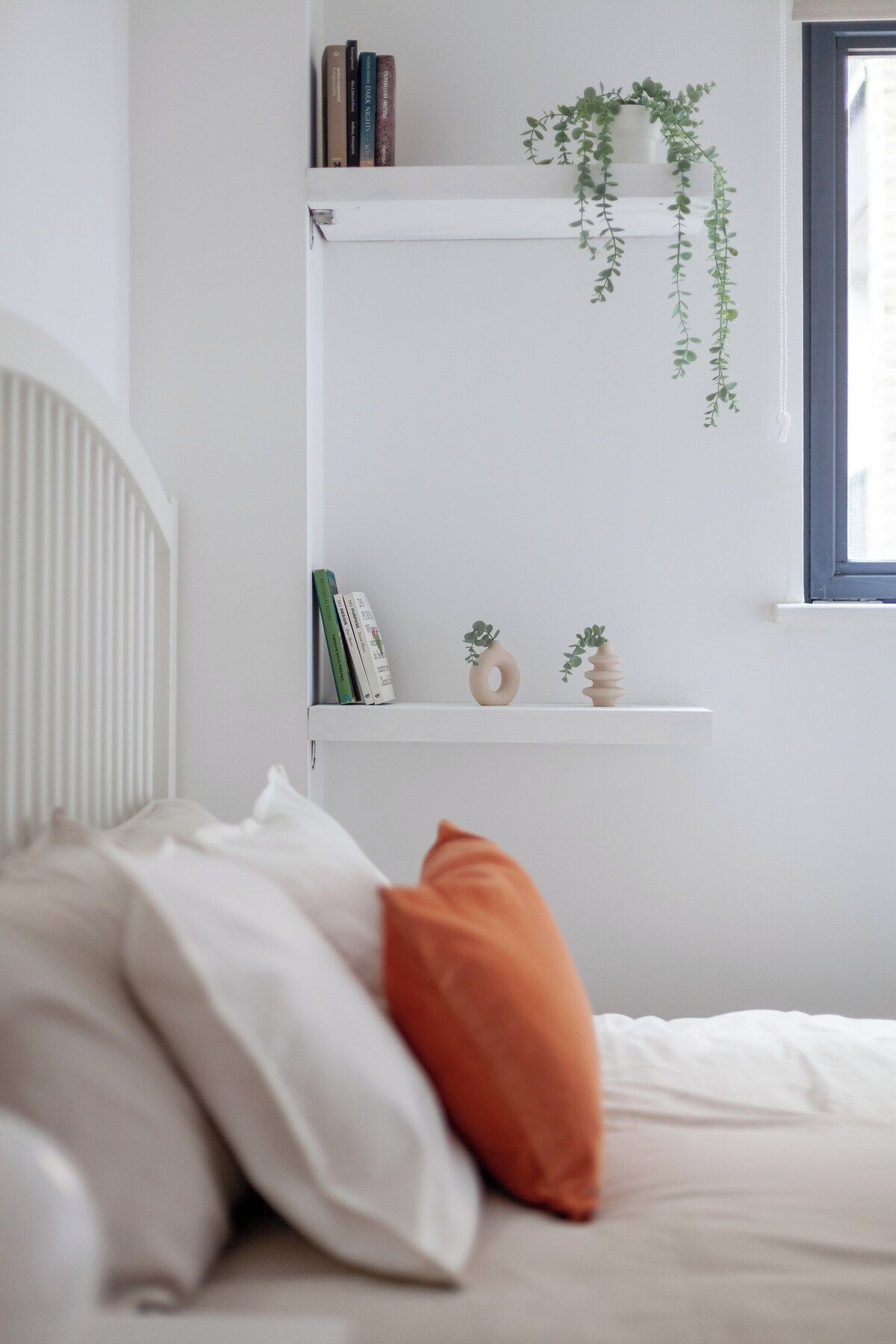 Ein Schlafzimmer mit einem Bett, Kissen, einem Fenster und Regalen mit Büchern und Pflanzen.