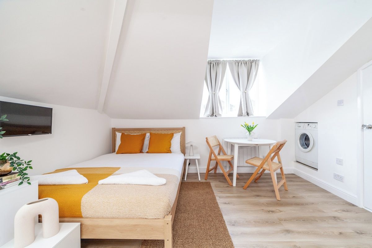 Un dormitorio con cama, escritorio, silla, ventana con cortinas, lavadora y televisor montado en la pared.
