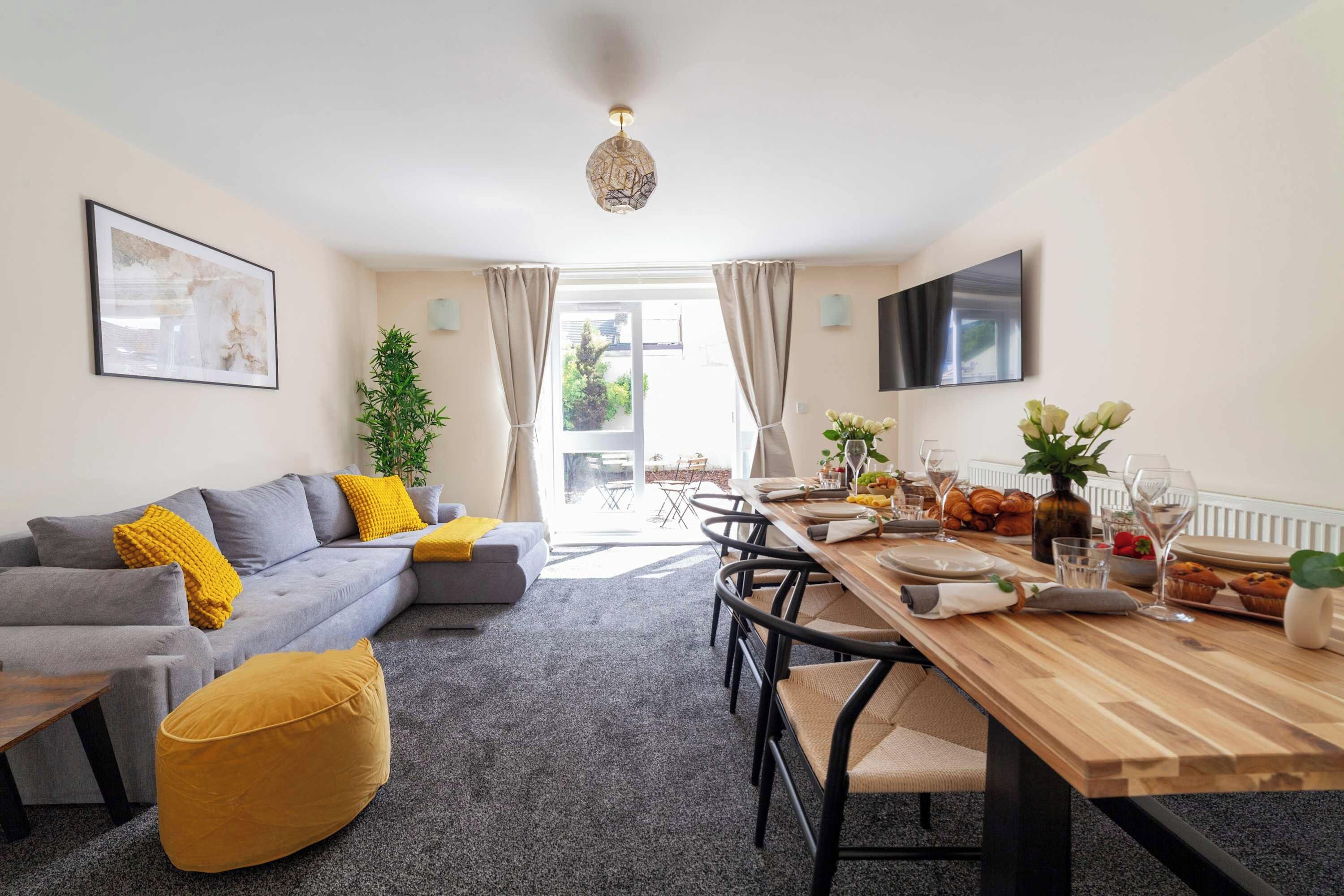 Un salon moderne avec un canapé gris, des coussins jaunes, une table à manger en bois mise pour le petit-déjeuner et un grand téléviseur fixé au mur.