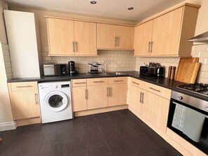 A modern kitchen with wooden cabinets, a washing machine, a dishwasher, a gas stove, and a coffee maker. - 3BR Stylish, Garden, Parking, Stratford (Greater London)