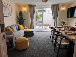 A modern living room with a grey sofa, yellow cushions, a round ottoman, a dining area with a wooden table set for a meal, and a large window with curtains. - 3BR Stylish, Garden, Parking, Stratford (Greater London)