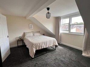 A bedroom with a bed, a bedside table, a lamp, a window with curtains, and a view of buildings outside. - 3BR Stylish, Garden, Parking, Stratford (Greater London)