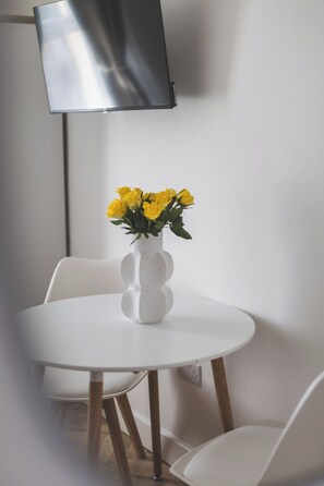 A white table with a white vase containing yellow flowers, a white chair, and a flat-screen TV mounted on the wall. - Stunning Studio Flat Highbury - GREAT Location (Greater London)