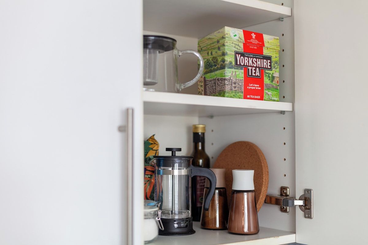 Un armario de cocina con una caja de Yorkshire Tea, una prensa francesa, un hervidor y un posavasos de corcho.