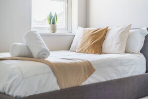 Ein ordentlich bezogenes Bett mit weißer Bettwäsche und einer beige farbenen Tagesdecke, ein Fenster mit einer Pflanze und ein grauer Kopfteil.