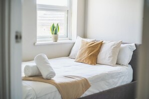 A neatly made bed with white bedding and a beige throw, a window with a potted plant, and a view of the outside. - 2 Bed| Sleeps 6|2 min Central line tube|Abba Arena (Greater London)