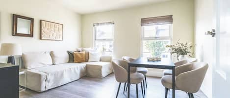 A modern living room with a sofa, dining table, and chairs, a window with a view, and artwork on the wall.