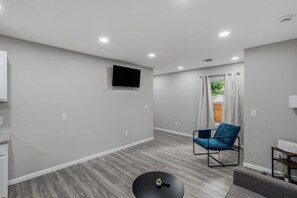 Ein modernes Wohnzimmer mit einem Wand-Fernseher, einem blauen Sessel, einem kleinen runden Couchtisch und Blick ins Freie durch ein Fenster mit Vorhängen.