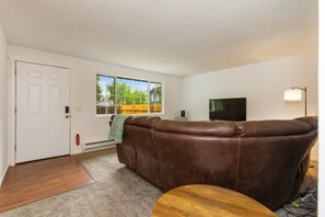 A living room with a brown leather sofa, a wooden coffee table, a flat-screen TV, and a window with a view of a fence and trees. - Cozy 2BR Home in Lynnwood w Backyard (Lynnwood)