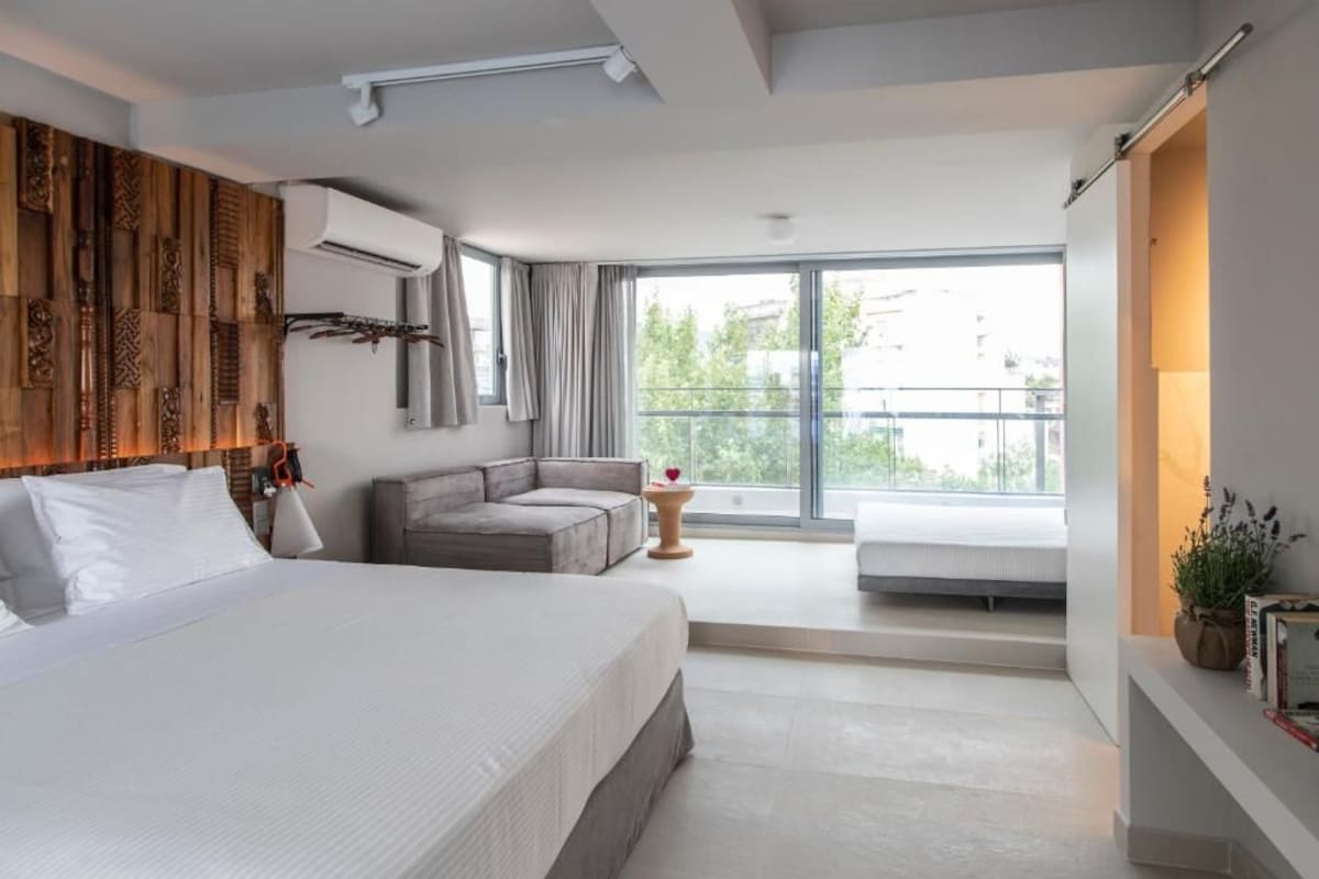 A modern hotel room with a large bed, a sofa, a small table, and a balcony with a view.