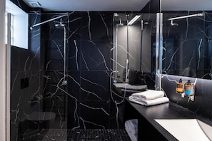 A modern bathroom with a black marble wall, a glass shower enclosure, and a vanity with a mirror and towel rack. - Citylinkers Suites (Athina)