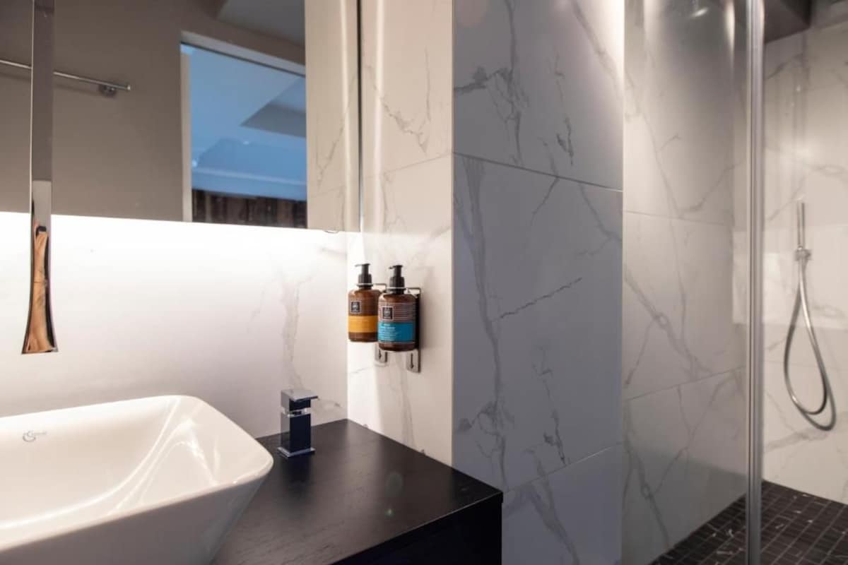 A modern bathroom with a large white sink, a dark countertop, marble-patterned walls, and a glass shower enclosure.