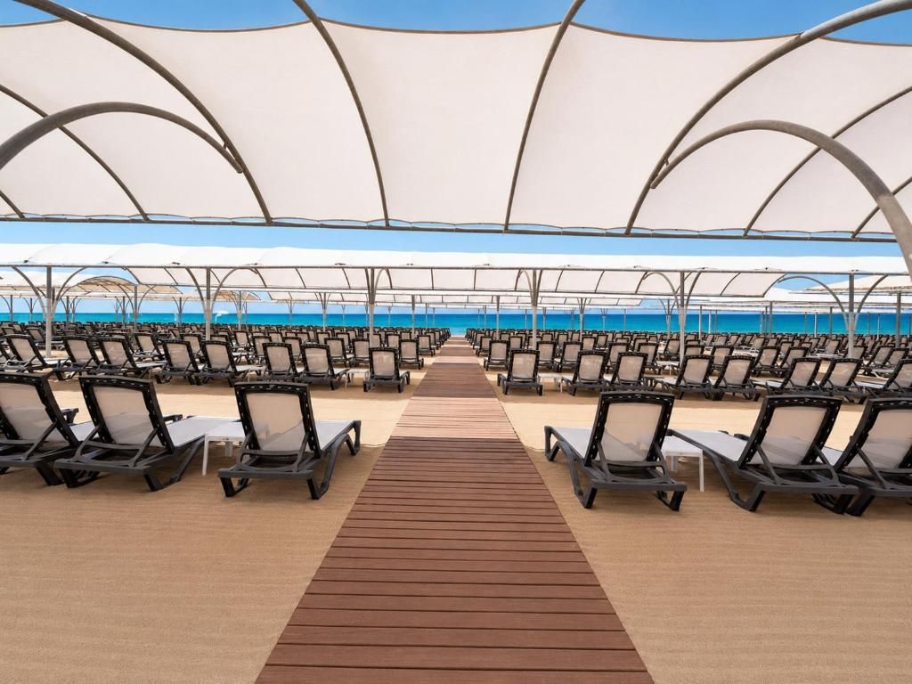 Un espacio de descanso frente a la playa, con filas de sillones reclinables y un paseo de madera que conduce al agua.