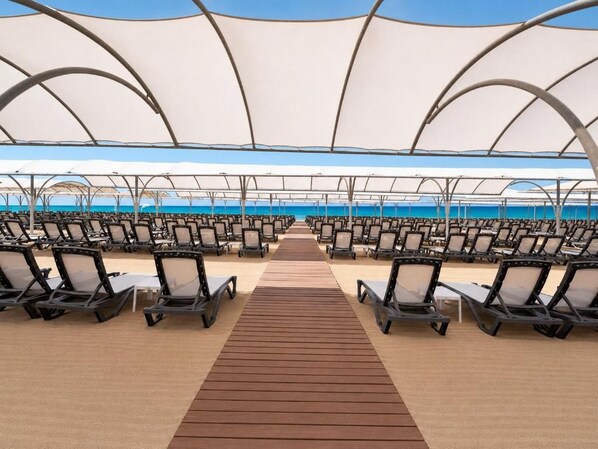 Un espacio de descanso frente a la playa, con filas de sillones reclinables y un paseo de madera que conduce al agua.