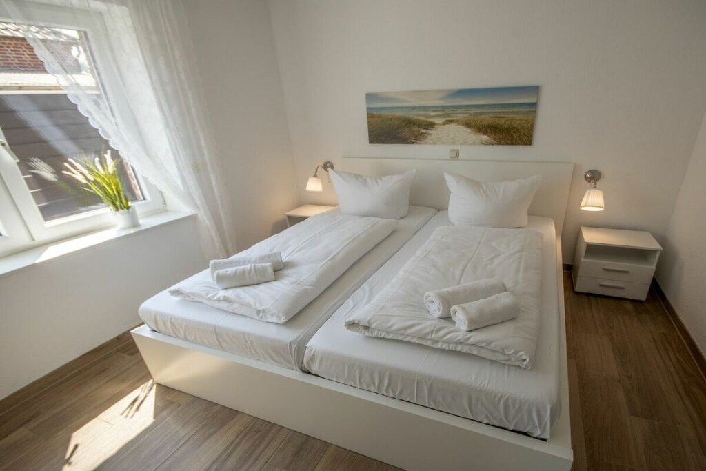 A double bed with white linens, two bedside tables with lamps, a window with curtains, and a painting on the wall.