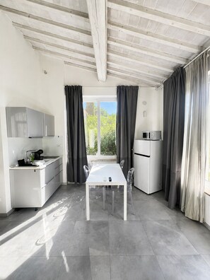 A modern kitchen with a white countertop, a built-in microwave, and a refrigerator. There is a window with curtains, a table with chairs, and a ceiling with exposed beams.