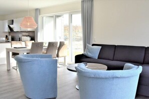 Ein modernes Wohnzimmer mit einem dunklen Sofa, hellen Sesseln, einem runden Couchtisch und einem Essbereich mit weißem Tisch und Stühlen.