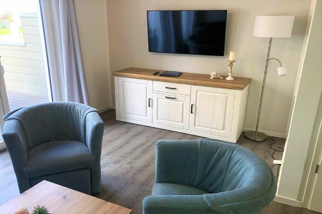 Ett rum med en tv som är monterad på väggen, ett vitt skåp med lådor, två grå fåtöljer och ett litet bord med en växt.