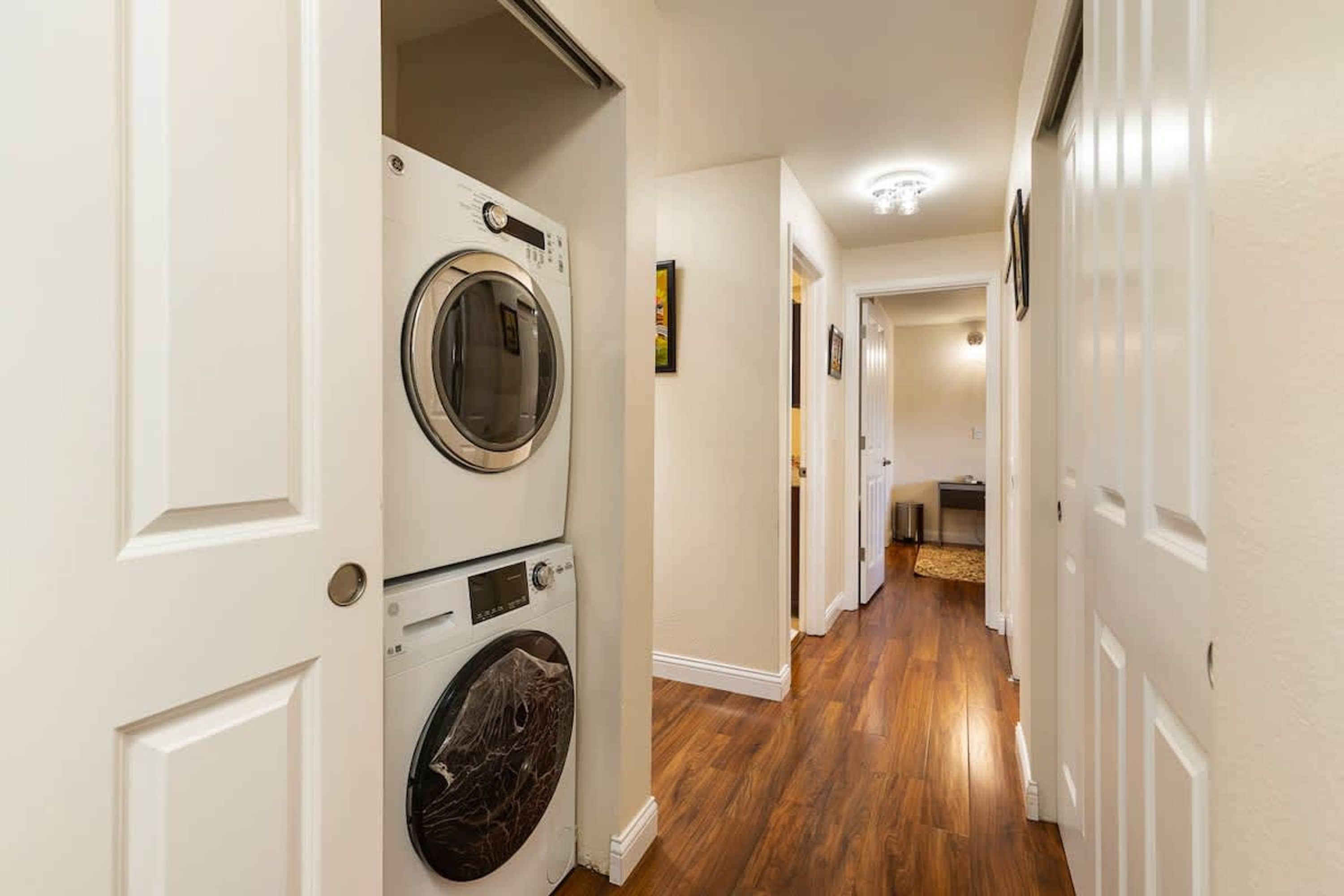 Une buanderie équipée d’une machine à laver et d’un sèche-linge superposés, avec un plancher en bois et un couloir menant à d’autres pièces.
