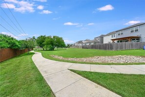 A residential backyard with a curved concrete pathway, a grassy lawn, a wooden fence, and houses in the background. - Spacious 2BR Flex Room Home Near Tech Campuses (Pflugerville)