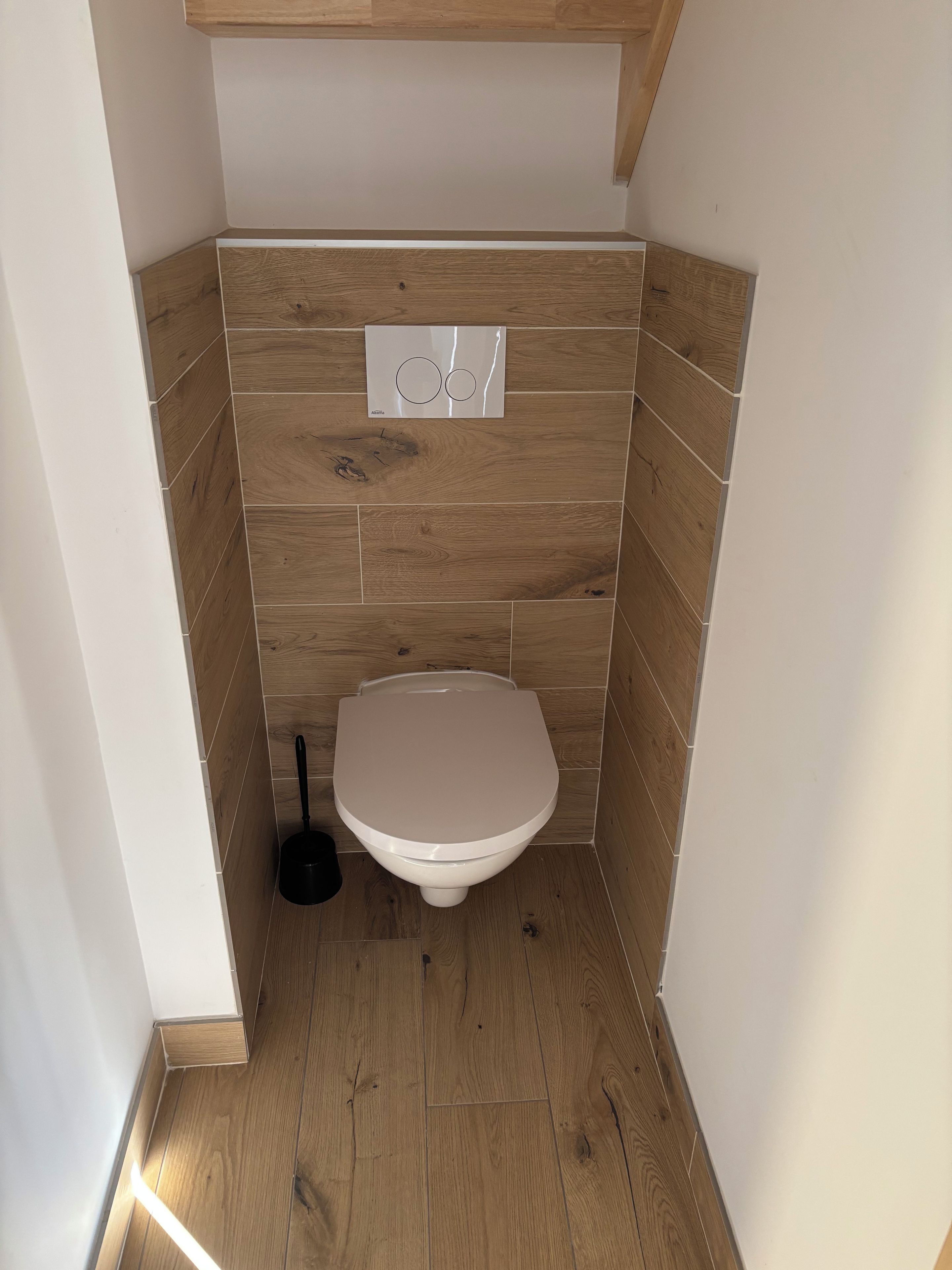Un baño pequeño y funcional con inodoro, una herramienta de limpieza negra y piso de madera.
