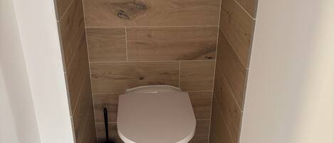 Un baño pequeño y funcional con inodoro, una herramienta de limpieza negra y piso de madera.