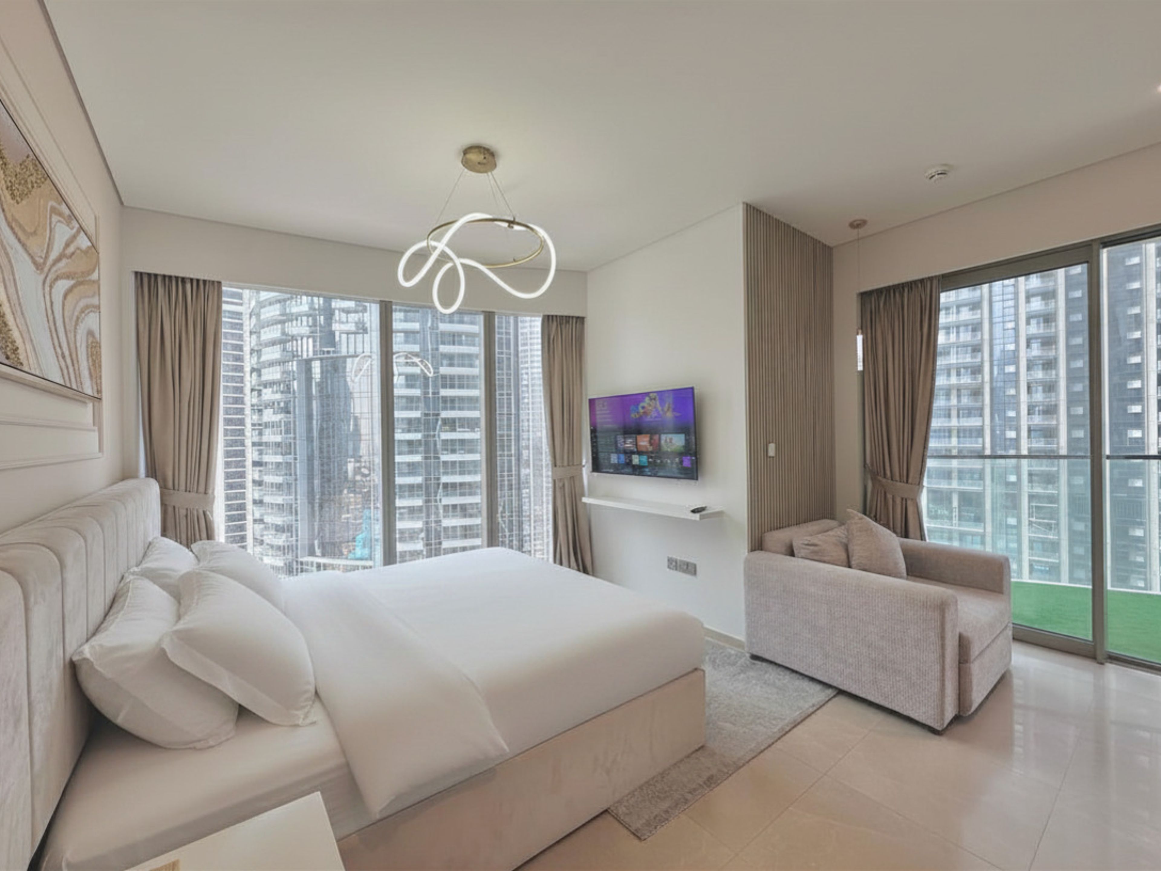A modern bedroom with a large bed, a TV mounted on the wall, and a view of high-rise buildings through the windows.