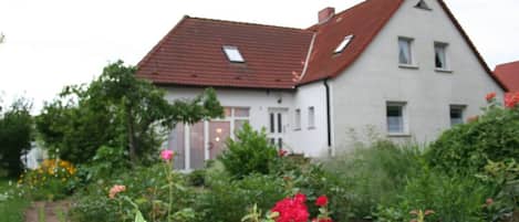 Una casa de dos pisos con techo rojo y paredes blancas, rodeada por un jardín con diversas plantas y flores.