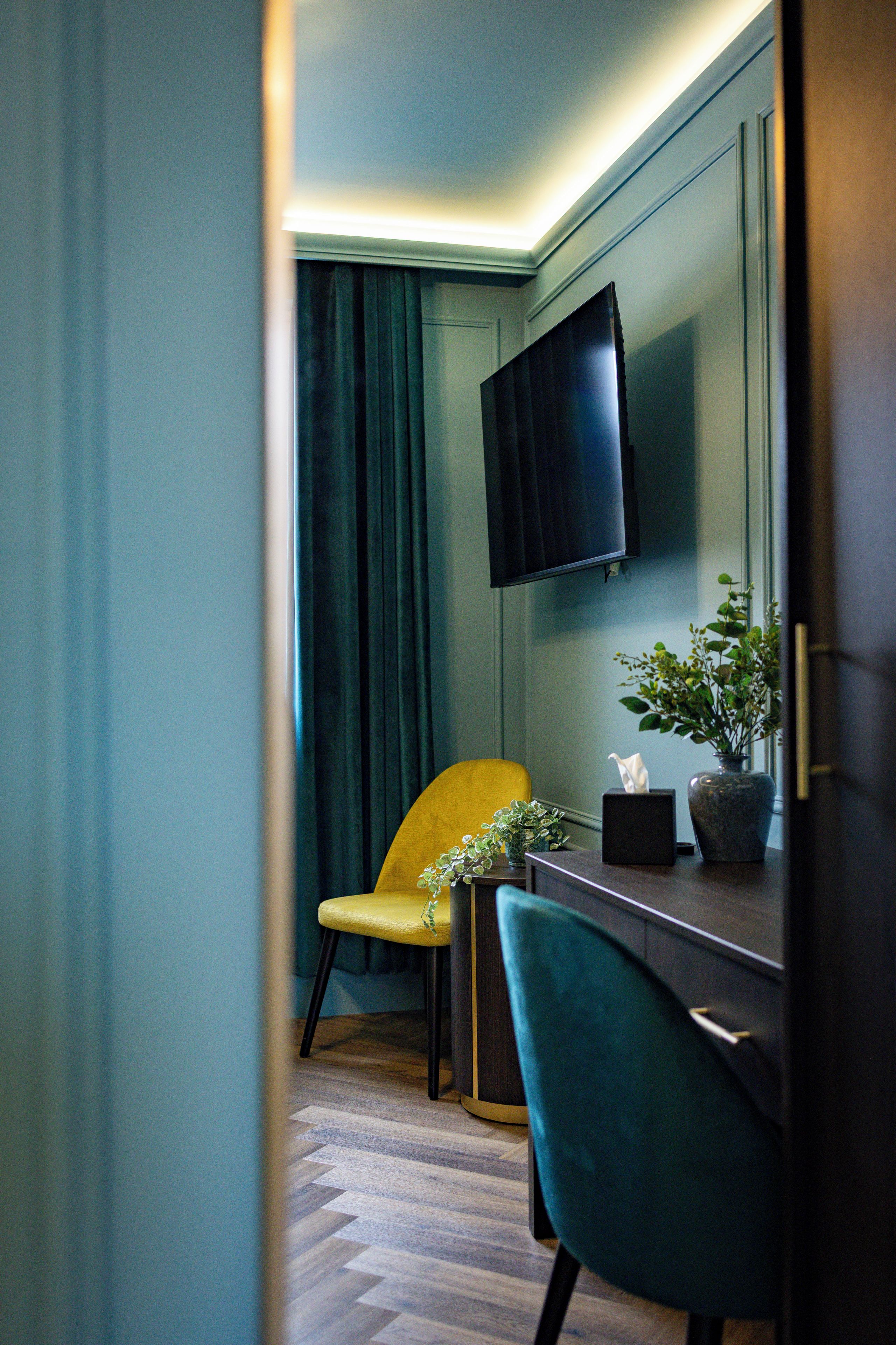 A room with a flat-screen TV mounted on the wall, a yellow chair, a teal chair, a small table with a plant, and a dark curtain.