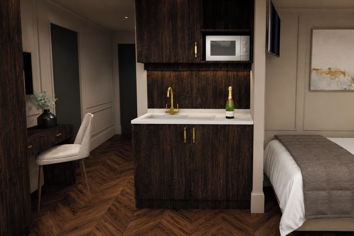 A compact hotel room with a built-in kitchenette, a bed with a gray blanket, a wooden herringbone floor, and a wall-mounted TV.