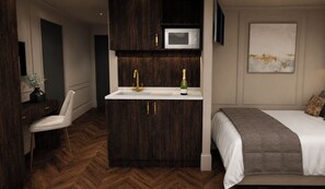 A compact hotel room with a built-in kitchenette, a bed with a gray blanket, a wooden herringbone floor, and a wall-mounted TV.