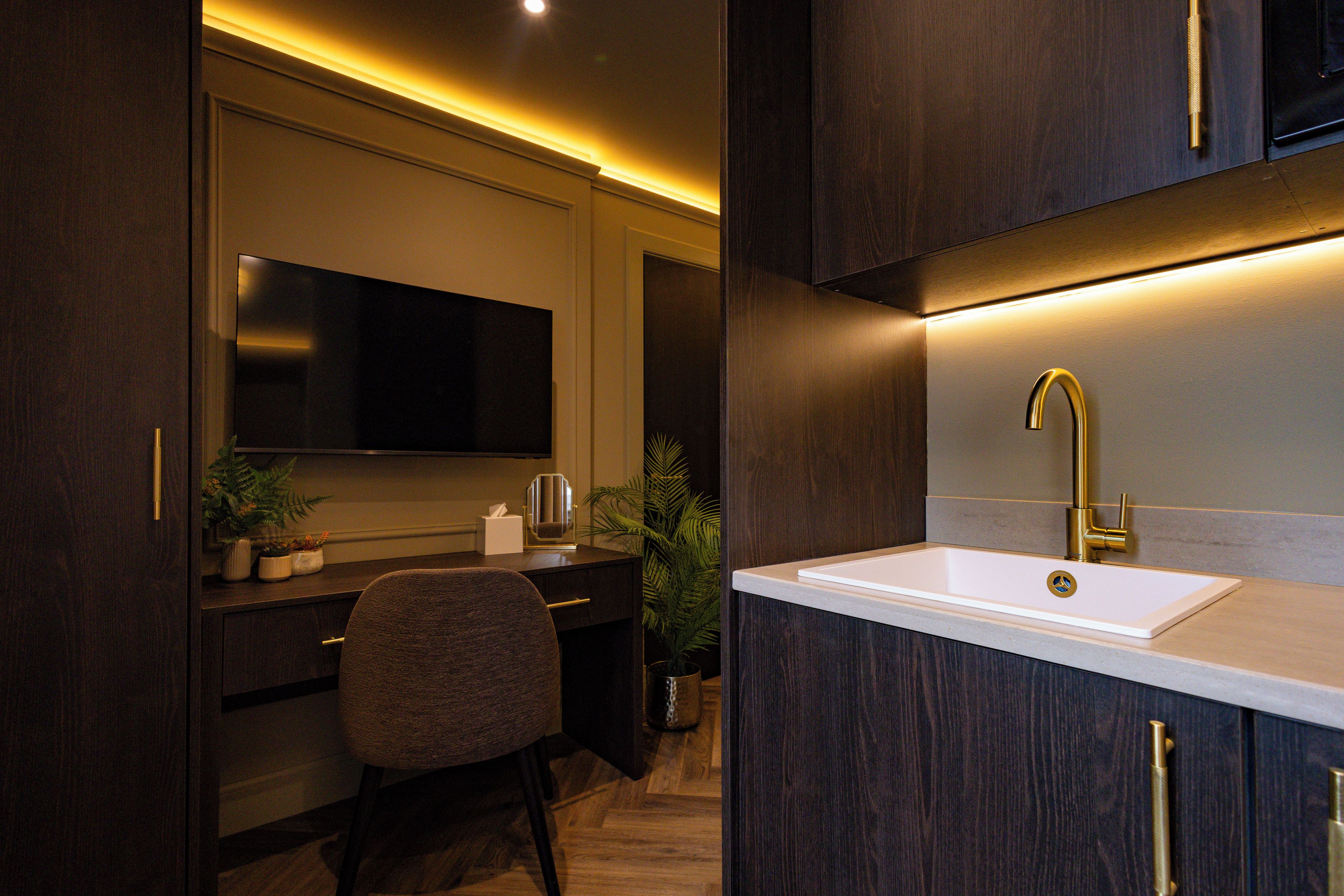 Eine moderne Küche mit integriertem Fernseher, einem weißen Spülbecken mit goldfarbenem Wasserhahn und dunklen Holzschränken.