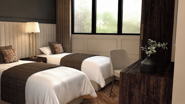 Ein Hotelzimmer mit zwei Betten, einem Nachttisch mit Lampe, einem Sessel und einem Fenster mit Blick auf Bäume.