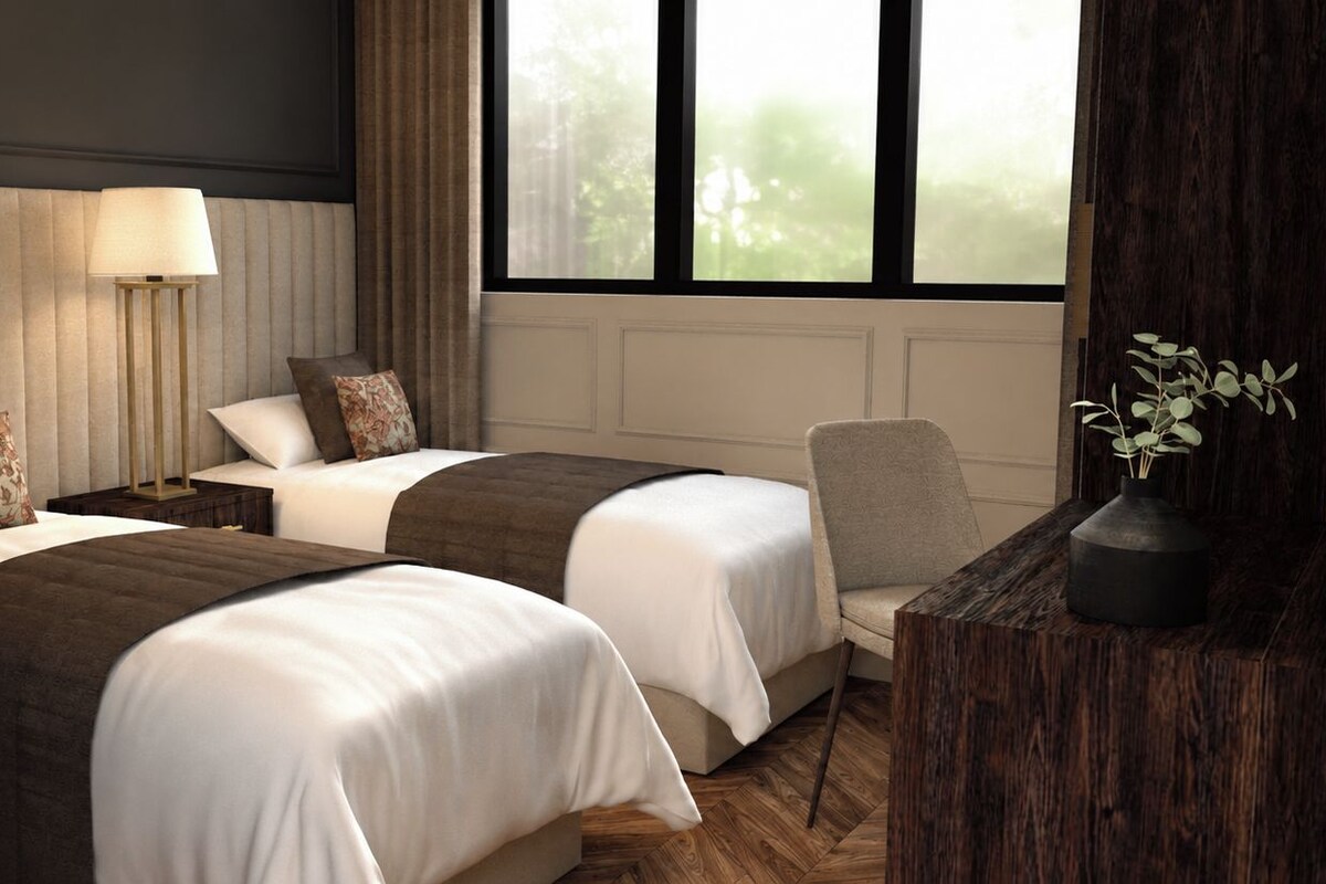 A hotel room with two beds, a bedside table with a lamp, a chair, and a window with a view of trees.