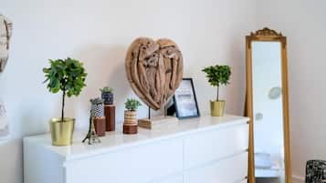 Un aparador blanco con objetos decorativos, incluyendo una escultura con forma de corazón, pequeñas macetas y una fotografía enmarco. Un espejo con marco dorado está a la derecha.