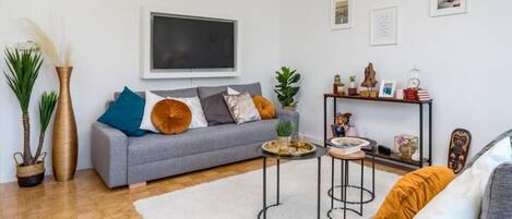 A modern living room with a grey sofa, a flat-screen TV, a small coffee table, and a bookshelf with various items.