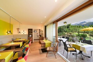 A dining area with multiple tables and chairs, a kitchen in the background, and a view of a garden and mountains through large windows. - House Rosengarten A&B" (Dorf Tirol)